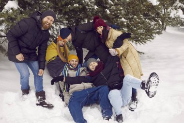 Funny cheerful young people having fun and enjoying snow in winter forest or park. Joyful men and women laughing loudly fall into snow. Concept of friendship and active life.