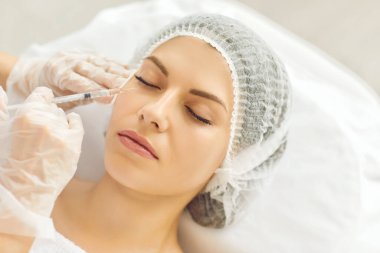 Young lady receives filler injection in lower eye lid area done by specialist at beauty clinic. Closeup womans head in medical cap, hands in gloves holding syringe. Aesthetic facial treatment concept