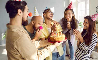 Smiling diverse people in festive cones congratulate excited girl with birthday present cake with candles. Happy friends greet overjoyed millennial colleague with anniversary. Celebration concept.