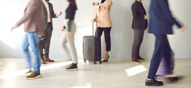 Woman with a suitcase waiting for transport and looking at her watch while standing at the train station or airport among a blurred crowd of people walking to and fro. Banner. Cropped shot, human legs