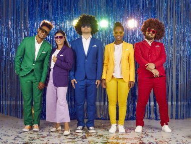 Portrait of young cheerful friends posing at theme party against shiny blue foil curtain. Multiracial people in colorful clothes, sunglasses and wigs stand in row and smile at camera. Full length.