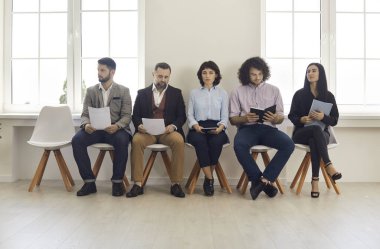 Group of five people sitting in office, holding documents and preparing for formal meeting or business appointment. Male and female candidates with CVs and resumes waiting in line for job interview