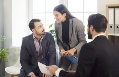 Group of a young business people sitting on workplace with documents discussing work project and company finance during a meeting. Coworkers and employees in formal clothes talking in office.