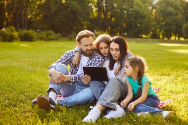 Happy family using tablet, sitting together on green grass of summer park. Mom and dad, kids hugging while reading digital book or text on modern device screen. Family picnic, bonding concept