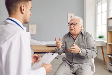 Yaşlı, beyaz saçlı bir adam muayenesi sırasında klinikte bir erkek doktorla konuşuyor. Yaşlı hastaların şikayetlerini dinleyen bir erkek doktor. Sağlık ve Tıp Konsepti.