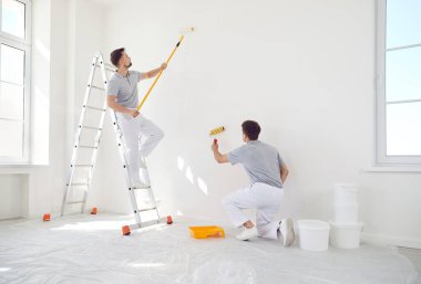 Two male painters in uniform paint a wall white with a roller. Home renovation concept. Professional work of builders. Modern apartment renovation, living room interior decoration