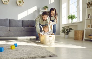 Young happy family having fun in the living room at home together with their child riding her in toy basket. Smiling couple spending time with a cute little daughter playing together.