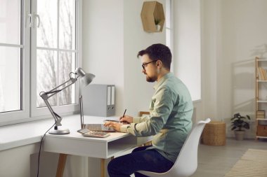 Attractive confident young man is sitting at his desk at home in the living room. Guy works remotely using laptop, taking notes by hand in notebook. Entrepreneur, accountant, engineer or freelancer.