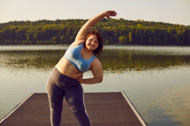 Happy fat young woman in sportswear doing stretching sport exercises in the summer city park on pier. Overweight girl exercising outdoors looking at camera. Body positive and outdoor training concept