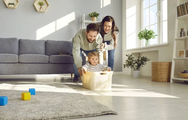 Young happy family having fun in the living room at home together with their child riding her in toy basket. Smiling couple spending time with a cute little daughter playing together.