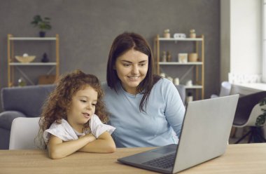 Happy mom and child using laptop at home. Mother and cute little daughter sitting at desk, looking at computer screen, video calling relatives or choosing good film, cartoon or family TV show to enjoy