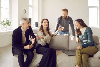 Group of happy coworkers having fun during the break at work. Four cheerful people of different ages gather on a comfortable sofa in a cozy, light office interior, sharing funny stories and laughing