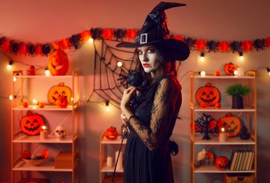 Portrait of mysterious and serious woman witch in room with Halloween decorations. Woman in black dress and witchs hat stands with black rose in her hands and looks at camera with terrible look.