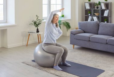 Old senior woman actively exercising indoors at home, sitting on large fitness ball, stretching, raising one arm bending, working on flexibility, fitness activity, elder enjoying workout at home
