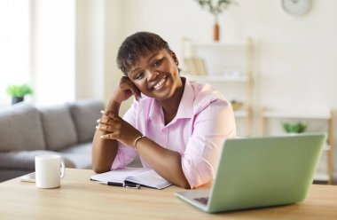 Portrait of a happy African American woman working on a laptop at her home office. She is smiling while sitting at a table, combining business tasks with the comfort of domestic living space.