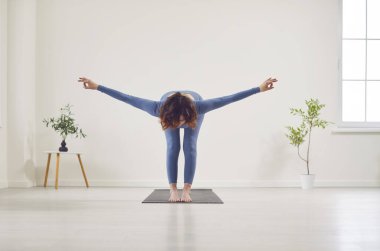 Attractive yogi woman working out, beautiful model doing yoga exercise on mat, standing in Ardha Uttanasana, Half Forward Bend pose, in home room or white studio among indoor green houseplants
