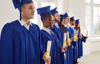 Group of happy smiling diverse multi ethnic college or university graduate students wearing traditional blue caps and gowns holding paper diploma scrolls standing in row at indoor graduation ceremony