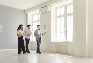 Portrait of young confident man professional realtor standing in empty house with couple clients showing new apartments. Man and woman buying property. Move, rent and real estate market concept.