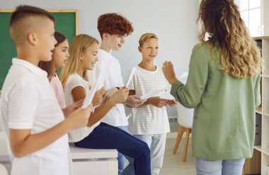 Group of happy smiling junior school children boys and girls standing in the classroom holding white sheets in hands with their young female teacher and singing in choir during music lesson.