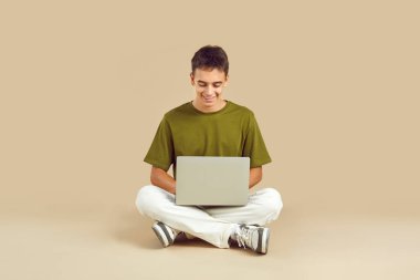 Portrait of a young smiling boy student wearing casual clothes working with laptop computer sitting in lotus pose isolated on a studio beige background. Education or remote work concept.