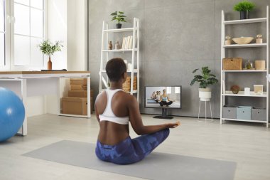 Back view of fit black woman sitting cross legged on yoga mat at home, having online video yoga class, looking at computer screen, learning to mediate, doing breathing hatha exercise, restoring energy
