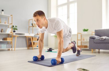 Strong, motivated, determined, young man in sportswear having a fitness workout at home, holding dumbbells, and doing a plank exercise with knee tucks on a rubber sports mat