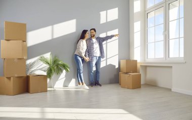 Loving husband and wife looking through window in their new home. Happy family couple standing in empty sunny living room with stacked cardboard boxes. Apartment ownership and rent, real estate