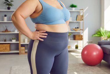 Cropped close up portrait of a belly and waist of fat overweight woman wearing sportswear standing at home after sport fit exercises. Workout, fitness, body positive and home training concept.