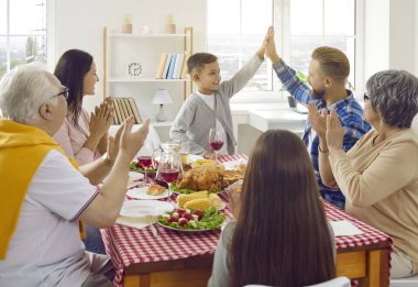 Joyful father and son give each other high five during family Thanksgiving dinner at home. Cheerful multigenerational family sits at festive table and applauds little boy. Family celebration concept.