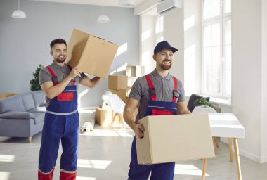 Relocation service. Loaders of moving and delivery company take out cardboard boxes from living room. Two smiling young male transportation professional workers in overalls help client with move