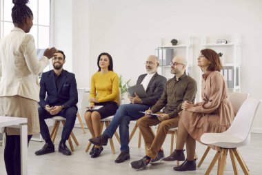 Female business coach speaks in front of small group of business people teaching them new business knowledge. Satisfied different people sitting on chairs in row and listening to dark skinned woman.