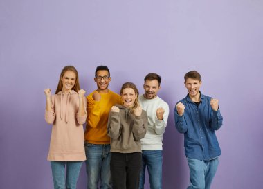 Victory and achievement. Happy positive young multiracial men and women rejoice together on purple background. Joyful people in casual clothes happily clench their fists looking at camera. Banner.
