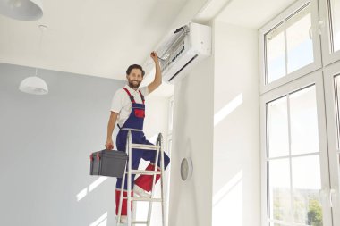 Profesyonel elektrikçi ya da teknisyen portresi merdivenin üzerinde duran, kameraya bakan ve gülümseyen, bakımlı, temizleyen, tamir eden ya da modern klima takan.