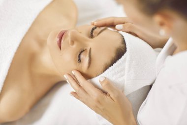 Close up of female masseur hands making face relaxing massage to young girl lying with closed eyes in beauty salon or cosmetology cabinet. Professional cosmetologist making massage for woman in spa.