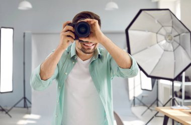 Smiling male professional photographer working in photo studio. Portrait of handsome young man taking photo with professional digital camera in studio with professional photographic equipment
