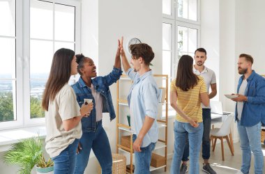 A group of young business people in elegant casual clothes communicate while standing in the office. Two cheerful young businessmen giving high five while their colleagues look at them and smile.