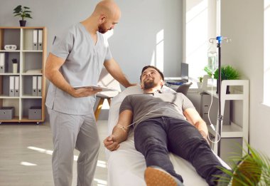 Young man lying on the couch at office in clinic and talking with a doctor while receiving IV drip infusion or vitamin therapy in his blood. Sick male person receiving injection therapy.