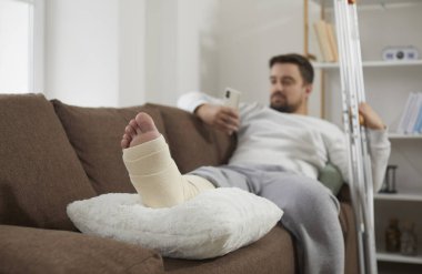 Man with a broken leg using his mobile phone while relaxing on the sofa at home, with his foot resting on a soft cushion pillow. Foot close up. Accident, injury, treatment, rehabilitation concept
