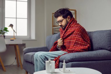 Sick young man in a warm blanket sitting on the sofa at home, feeling cold, freezing, shivering, drinking hot tea and taking medicine. Concept of seasonal cold and flu infection