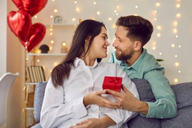 Happy, handsome, young man gives gift to his attractive lover on Valentines day, birthday, holiday. Young couple in love in romantic atmosphere are sitting on sofa in cozy living room at home.