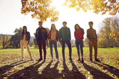 Six confident young people in casual outfits enjoying leisure, posing in park on sunny autumn day, standing back lit, casting shadows on green grass and yellow leaves. Full body backlight group shot