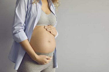 Pregnant belly. Close up of naked belly of pregnant woman gently stroking it and touching posing on gray background. Woman in third trimester of pregnancy in comfortable clothes near copy space.