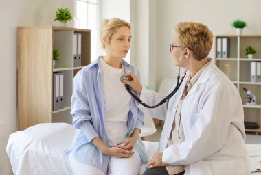Doctor using stethoscope to perform chest auscultation for young female patient sitting on exam bed in clinic and breathing. Professional physician listening and thinking of girls diagnosis