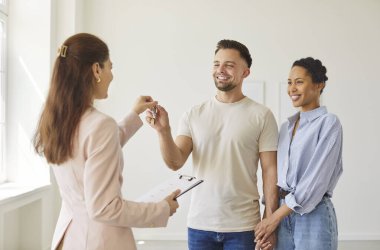 Happy homeowners getting keys of their new apartment from real estate agent, ready to start new life in new house. Woman realtor congratulating family couple with purchasing dwelling