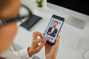 Closeup of woman looking at phone display with red like heart and message buttons under photo of attractive young man on online dating website or mobile phone app. Hands holding smartphone in close up