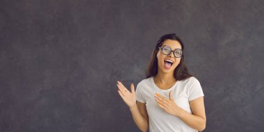Young funny comic woman isolated on gray studio background pointing at blank copy space aside. Caucasian casual brunette woman in white t-shirt and funny glasses having fun advertising. Banner.