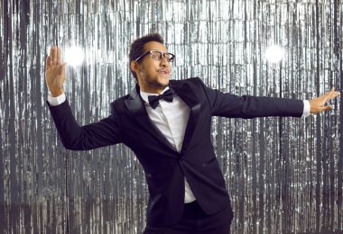 Positive young african american man in elegant suit having fun dancing at party on shiny background. Guy in glasses, tuxedo and bow tie dances with funny expression on silver background.