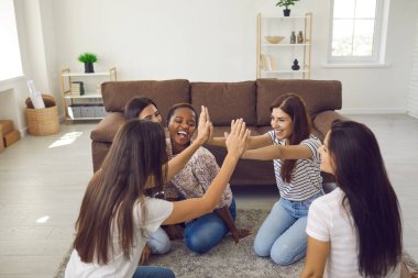 Happy excited best friends high fiving each other sitting on floor at home. Diverse group of positive cheerful confident young women having fun together at casual gathering in friends apartment