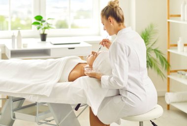Young woman cosmetologist applying moisturizing mask with brush to female pretty client lying on the couch in beauty spa salon. Girl receiving beauty skin care cosmetology procedure from beautician.