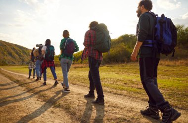 Group of people on a trekking or hiking tour. Back view several male and female hikers with backpacks walking on a dirt trail among green trees and hills under sunny sky. Tourism concept